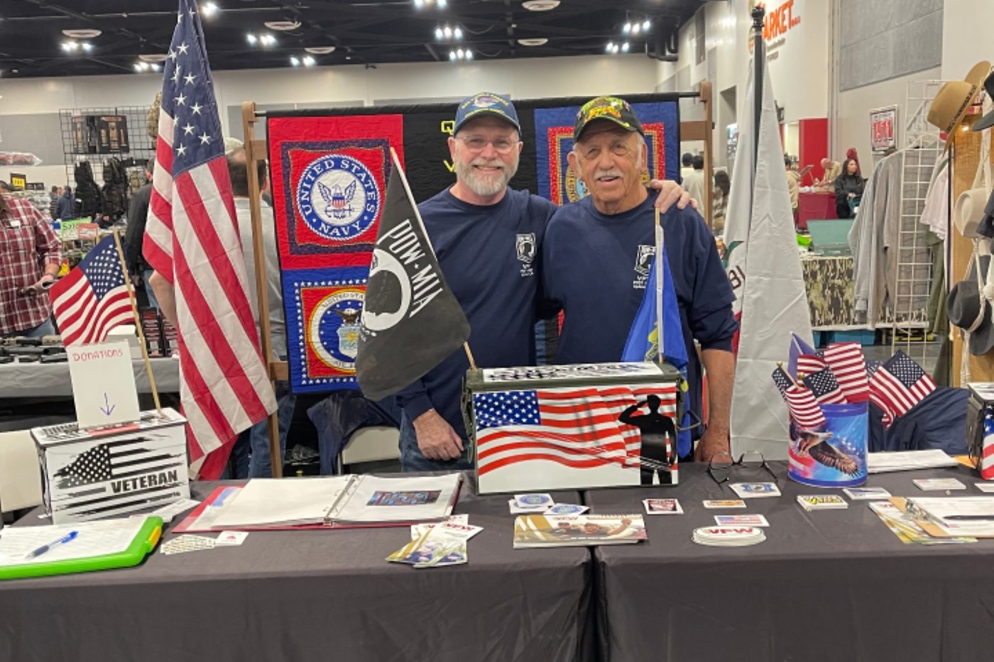 Two VFW Post 10489 members at an indoor outreach table with American flags and informational materials